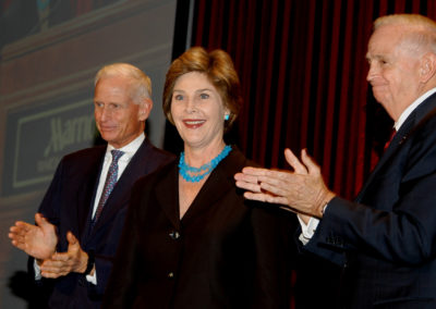 First Lady Laura Bush smiling in a dark dress with blue necklace and earrings flanked by Richard E. Marriott clapping to her left and J.W. “Bill” Marriott, Jr. clapping to her right at the 2005 Bridges Gala
