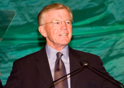 Former Washington Redskins Head Coach Joe Gibbs in a dark suit smiling at a podium with a green backdrop at the 2006 Bridges Gala