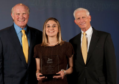 Smiling young female participant holding an award with smiling NFL legend Terry Bradshaw in a dark suit with yellow tile standing to her left and smiling Richard E. Marriott standing to her right at the 2010 Bridges Gala